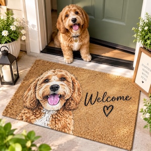 Puede incluir: Un felpudo rectangular de fibra de coco color canela con una ilustración impresa de un perro de pelo rizado color marrón dorado con la lengua fuera. El texto "Welcome" está impreso en cursiva negra junto a un pequeño corazón negro.