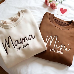 Könnte beinhalten: Zwei Sweatshirts mit Rundhalsausschnitt, eines cremefarbenes und eines braunes. Das cremefarbene Sweatshirt hat das Wort "Mama" in Dunkelbraun gestickt, darunter "EST. 2026". Das braune Sweatshirt hat "Mini" in Weiß gestickt, darunter "EST. 2026".