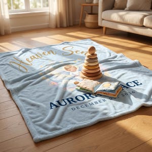K&ouml;nnte beinhalten: Hellblaue Babydecke mit goldfarbenem Schriftzug "Heaven Sent" und dem Namen "Aurora" in Marineblau. Ein Holz-Steckspielzeug und ein Stoffbuch liegen auf der Decke. Die Decke hat eine Engel-Illustration.