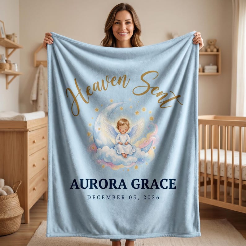 K&ouml;nnte beinhalten: Hellblaue Babydecke mit goldfarbenem Schriftzug "Heaven Sent". Die Decke zeigt eine Illustration eines Engels auf einem Mond, mit dem Namen "AURORA GRACE" und dem Datum "DECEMBER 05, 2026" darunter.