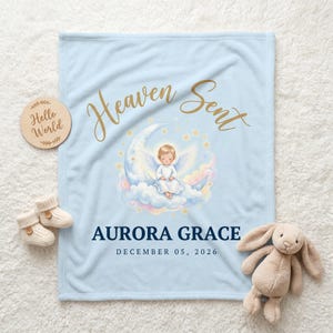 K&ouml;nnte beinhalten: Hellblaue Babydecke mit goldfarbenem Schriftzug "Heaven Sent". Die Decke zeigt eine Illustration eines Engels auf einem Mond. Der Name "Aurora Grace" und das Datum "05. Dezember 2026" sind darunter aufgedruckt.