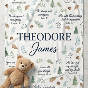 May include: A personalized baby blanket with the name "THEODORE JAMES" in navy blue script. The blanket features botanical illustrations, Bible verses, and a plush teddy bear. The blanket is white with a soft texture.