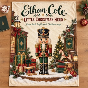 May include: A cream-colored fleece blanket featuring a central nutcracker figure, wrapped gifts, a rocking horse, and a teddy bear. The text reads Ethan Cole, LITTLE CHRISTMAS HERO, and Brave heart, bright spirit, Christmas magic in dark green and red.
