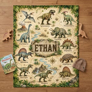 May include: A rectangular blanket featuring a prehistoric map design with illustrations of various dinosaurs including a Tyrannosaurus, Triceratops, and Pterodactyl. The center displays the name ETHAN on a rock graphic with jungle foliage.