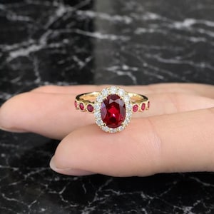 May include: A gold ring with a large oval ruby centre stone, encircled by a halo of small diamonds. The ring's band is decorated with small, round rubies. The ring is set against a dark background.