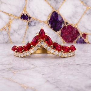 Könnte beinhalten: Ein goldener Ring mit V-förmigem Design, besetzt mit leuchtend roten, marquiseförmigen Edelsteinen und kleinen, funkelnden weißen Diamanten. Der Ring ist vor einem Marmorhintergrund mit goldenen und violetten Akzenten platziert.