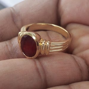 May include: A gold ring featuring a large, oval-cut, deep red gemstone. The ring has a smooth, polished band and decorative ribbed detailing on the sides of the gemstone setting. The ring is shown in a close-up view.