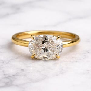 May include: A gold engagement ring with a large, oval-cut diamond. The diamond is held in place by four prongs, and the band is a simple, polished gold. The ring is set against a white background.