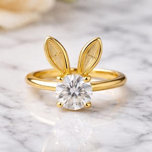 Puede incluir: Un anillo dorado con una gran gema redonda y transparente. El diseño del anillo incluye dos orejas de conejo doradas, colocadas sobre la gema. El fondo está borroso.