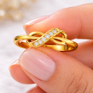 May include: A gold-colored infinity ring with a row of small, clear gemstones set across the top. The ring is held between two fingers, showcasing its polished finish and intricate design. The ring's design is a symbol of eternity.