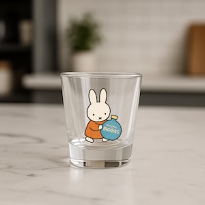 May include: A clear glass tumbler featuring a cartoon rabbit character in an orange dress, holding a blue bottle with the text "BuzzBallz Biggies". The glass is set on a white surface.