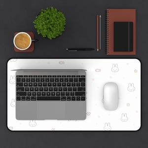 Pode incluir: Uma vista de cima de uma configuração de mesa com um laptop, um mouse e um mousepad branco com um padrão de coelhos e peixes. Uma xícara de café, uma pequena planta e artigos de papelaria também estão presentes. O fundo é cinza escuro.