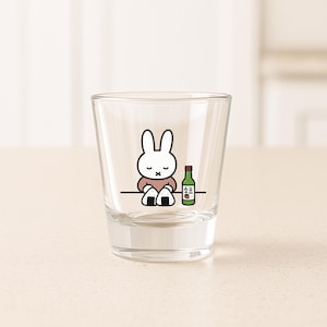 May include: A clear shot glass featuring a cartoon rabbit character with a pink shirt and a small bottle of soju. The rabbit is sitting with its legs crossed. The bottle is green with white text.