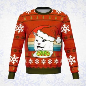 May include: A long-sleeve crewneck sweatshirt in bright orange featuring a repeating pattern of white snowflakes and green pine trees. The center graphic displays a white cat wearing a red Santa hat behind a plate of salad against a sunset background.