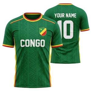 May include: Green football shirt with yellow and red detailing. The front features the word "CONGO" in white, and a shield emblem. The back has the number 10 and space for a name. The shirt has a patterned design.