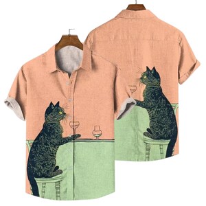 May include: A short-sleeved button-down shirt with a peach-colored front and back. The shirt features a whimsical illustration of a black cat sitting at a table with a glass of wine. The cat is depicted on both the front and back of the shirt.