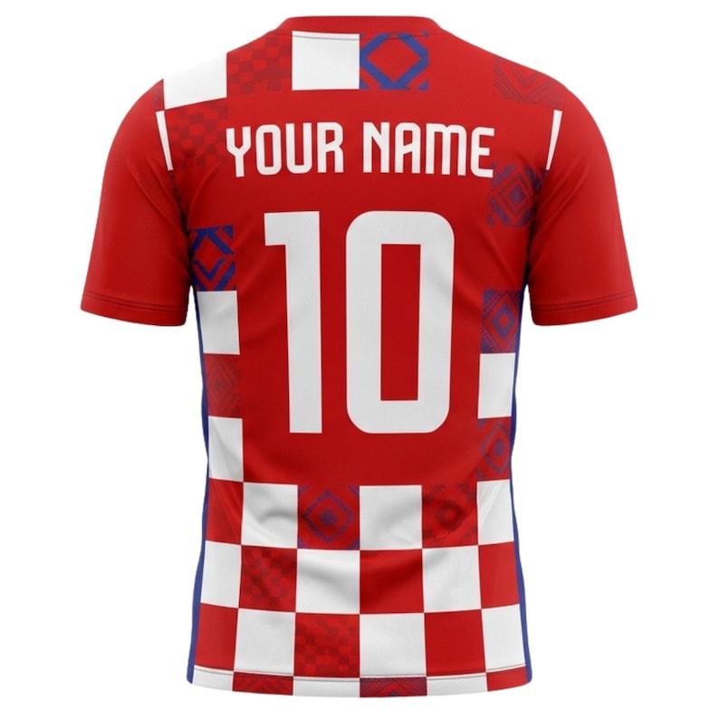 May include: Red football shirt with white and blue detailing. The back displays the text "YOUR NAME" above the number "10" in white. The shirt has a checkered pattern at the bottom and a design on the shoulders.