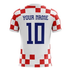 Op de afbeelding: Wit voetbalshirt met een rood-wit geblokt patroon op de schouders en mouwen. De achterkant toont het nummer 10 in blauw en de tekst "YOUR NAME". Een sportkledingartikel.