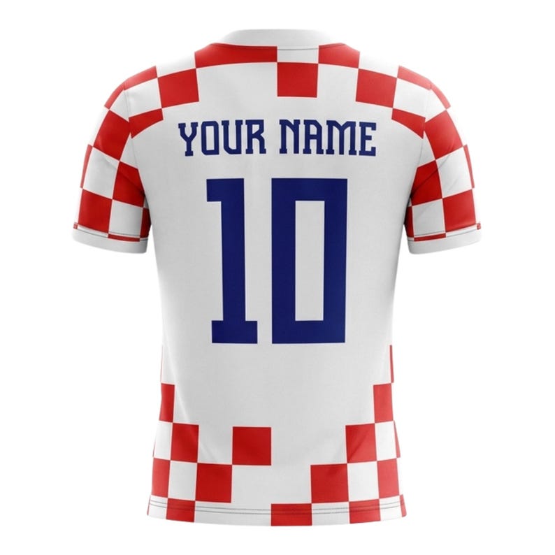 Op de afbeelding: Wit voetbalshirt met een rood-wit geblokt patroon op de schouders en mouwen. De achterkant toont het nummer 10 in blauw en de tekst "YOUR NAME". Een sportkledingartikel.