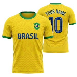 Puede incluir: Camiseta de f&uacute;tbol amarilla de Brasil con ribetes verdes y la palabra "BRASIL" en azul. La camiseta presenta la bandera brasile&ntilde;a y un estampado de hojas. La parte trasera de la camiseta tiene el n&uacute;mero 10 y el texto "YOUR NAME" en azul.