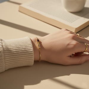 Puede incluir: Una delicada pulsera dorada con tres piedras preciosas rectangulares: una p&uacute;rpura oscura, una transparente y una amarillo p&aacute;lido. La pulsera est&aacute; en una mu&ntilde;eca, con una manga de su&eacute;ter color crema visible. Un anillo de oro est&aacute; en el dedo.
