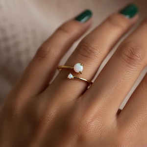 May include: A gold-colored ring with two white opal stones. The ring has a delicate, open design, with one larger opal and a smaller one. The ring is worn on a finger with a green nail polish.