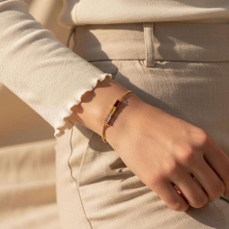 Puede incluir: Una pulsera dorada con una cadena delicada y tres piedras rectangulares: una transparente, una amarilla y una roja. La pulsera se lleva en la mu&ntilde;eca, con una camisa de manga larga color crema y pantalones color beige en el fondo.