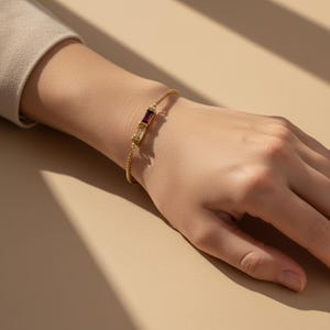 Puede incluir: Una pulsera dorada con una cadena delicada y una pieza central rectangular. La pieza central presenta una piedra preciosa de color rojo intenso y amarillo claro. La pulsera se lleva en una mu&ntilde;eca sobre un fondo neutro.