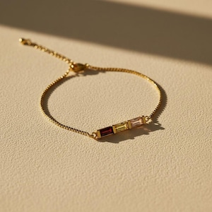 Puede incluir: Un brazalete dorado con una delicada cadena y tres piedras rectangulares en tonos rojo, amarillo y rosa. El brazalete se presenta sobre un fondo neutro, resaltando el dise&ntilde;o de la joya.