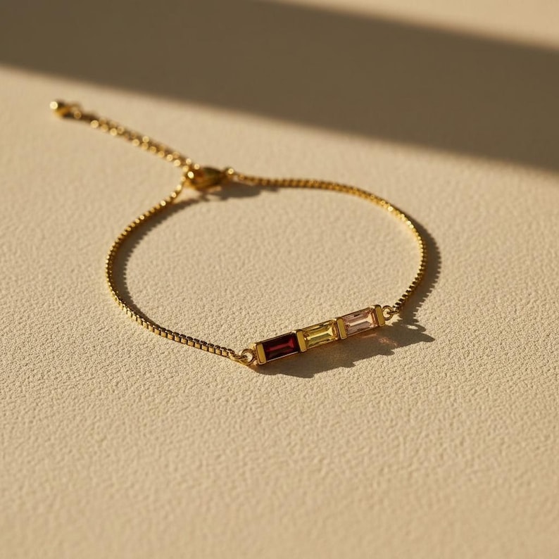Puede incluir: Un brazalete dorado con una delicada cadena y tres piedras rectangulares en tonos rojo, amarillo y rosa. El brazalete se presenta sobre un fondo neutro, resaltando el dise&ntilde;o de la joya.
