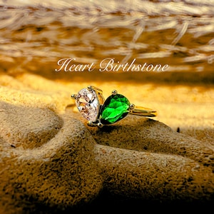 May include: A gold ring featuring two pear-shaped gemstones: one clear and one emerald green. The ring is set on a textured, tan surface. The text "Heart Birthstone" is visible in the image.