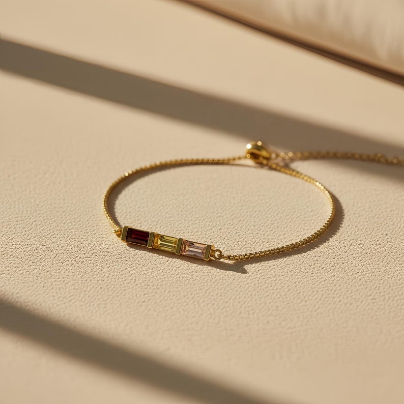 Puede incluir: Una pulsera dorada con tres gemas rectangulares en tonos rojo, amarillo y rosa. La pulsera tiene una cadena fina y un cierre deslizante ajustable. Las gemas est&aacute;n dispuestas horizontalmente.