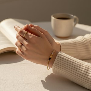 Puede incluir: Una pulsera dorada con piedras preciosas rectangulares en negro, transparente y amarillo se lleva en la mu&ntilde;eca. Dos anillos dorados est&aacute;n en los dedos. Una taza de caf&eacute; blanca y un libro abierto est&aacute;n en el fondo.