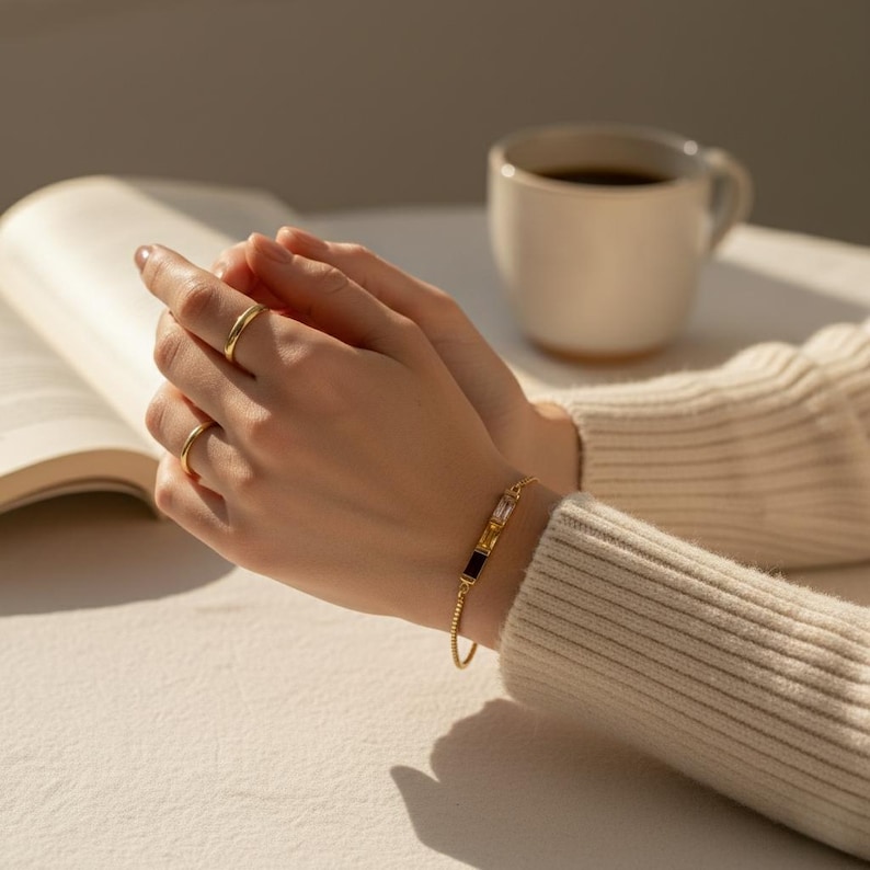 Puede incluir: Una pulsera dorada con piedras preciosas rectangulares en negro, transparente y amarillo se lleva en la mu&ntilde;eca. Dos anillos dorados est&aacute;n en los dedos. Una taza de caf&eacute; blanca y un libro abierto est&aacute;n en el fondo.