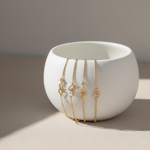 Puede incluir: Cuatro pulseras doradas con delicadas cadenas y diseños de abalorios únicos, incluyendo la palabra "love" en una fuente estilizada. Cada pulsera presenta un pequeño acento brillante. Las pulseras se exhiben dentro de un cuenco de cerámica blanco y redondeado.