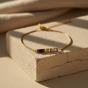 Puede incluir: Una pulsera dorada con tres piedras rectangulares en tonos rojo, amarillo y rosa. La pulsera est&aacute; sobre una superficie texturizada blanquecina, con un fondo suave y neutro.