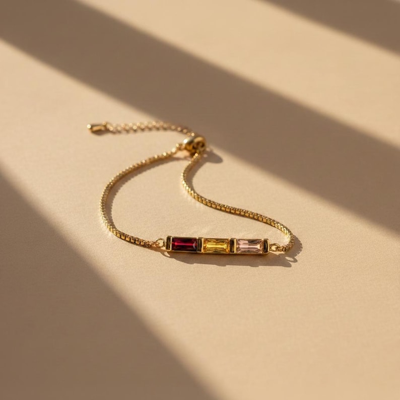 Puede incluir: Una pulsera dorada con una cadena fina y una barra rectangular con tres piedras preciosas de colores: un rojo intenso, un amarillo brillante y un rosa suave. La pulsera se muestra sobre un fondo neutro.