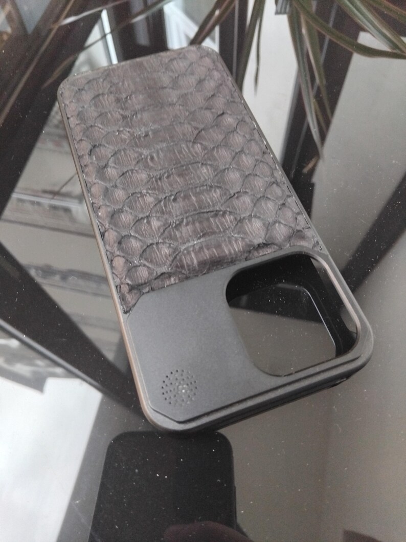 Pode incluir: Uma capa de telefone cinza escuro com um padr&atilde;o texturizado em forma de escamas na parte superior. A parte inferior &eacute; preta fosca s&oacute;lida com recortes para a c&acirc;mera e o alto-falante. A capa tem um design elegante e moderno.