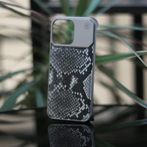 Op de afbeelding: Smartphonehoesje met een slangenprint in grijstinten en wit. De hoes heeft een zilverkleurig frame en een camera-uitsparing. Ontworpen om de telefoon te beschermen en een stijlvolle uitstraling te geven.