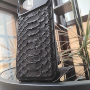 Pode incluir: Capa de telefone preta com um padr&atilde;o texturizado em forma de escamas. A capa tem um acabamento fosco e um protetor de lente de c&acirc;mera. O design lembra a pele de cobra ou r&eacute;ptil, oferecendo uma est&eacute;tica &uacute;nica. A capa foi projetada para proteger o telefone.