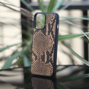 May include: A black phone case with a snakeskin pattern in shades of brown and tan. The case is standing upright, showcasing the design and texture. The background features green plants and a dark surface.