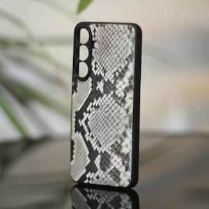 Puede incluir: Una funda para smartphone con borde negro y un estampado de piel de serpiente en tonos grises y blancos. La funda tiene recortes para la cámara y otras funciones del teléfono. La funda está de pie.