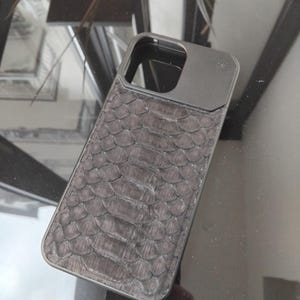 Pode incluir: Uma capa de telefone cinza escuro com um padr&atilde;o texturizado e escamoso, possivelmente imitando pele de cobra. A capa tem formato retangular com recorte para a lente da c&acirc;mera e uma moldura de metal escuro.