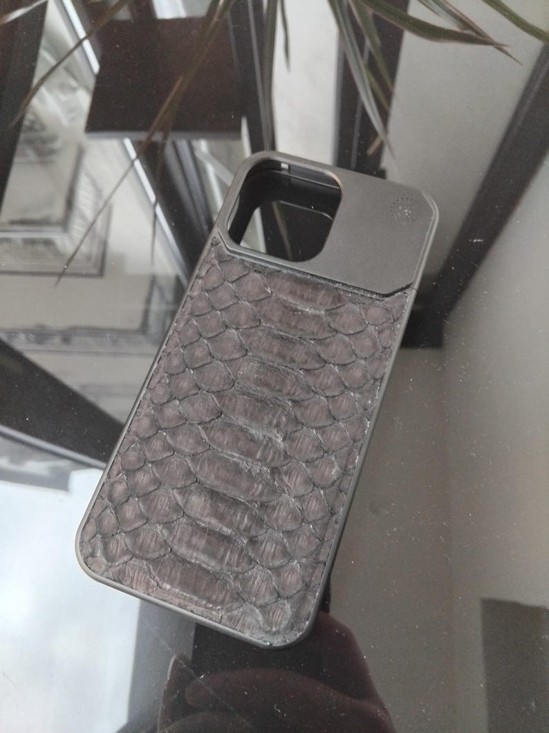 Pode incluir: Uma capa de telefone cinza escuro com um padr&atilde;o texturizado e escamoso, possivelmente imitando pele de cobra. A capa tem formato retangular com recorte para a lente da c&acirc;mera e uma moldura de metal escuro.