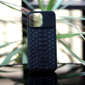 Pode incluir: Capa de telefone preta com um padr&atilde;o texturizado semelhante &agrave; pele de cobra. A capa tem um recorte para a lente da c&acirc;mera e &eacute; feita de um material escuro. A capa do telefone &eacute; exibida em uma superf&iacute;cie reflexiva com plantas verdes ao fundo.