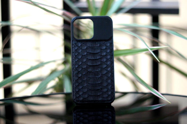 Pode incluir: Capa de telefone preta com um padr&atilde;o texturizado semelhante &agrave; pele de cobra. A capa tem um recorte para a lente da c&acirc;mera e &eacute; feita de um material escuro. A capa do telefone &eacute; exibida em uma superf&iacute;cie reflexiva com plantas verdes ao fundo.