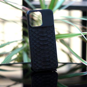 Pode incluir: Uma capa de telefone preta com um padr&atilde;o texturizado de pele de cobra. A capa tem um recorte para a c&acirc;mera e est&aacute; em um fundo desfocado de plantas verdes e uma superf&iacute;cie reflexiva. A capa foi projetada para proteger um smartphone.