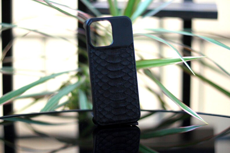 Pode incluir: Uma capa de telefone preta com um padr&atilde;o texturizado de pele de cobra. A capa tem um recorte para a c&acirc;mera e est&aacute; em um fundo desfocado de plantas verdes e uma superf&iacute;cie reflexiva. A capa foi projetada para proteger um smartphone.