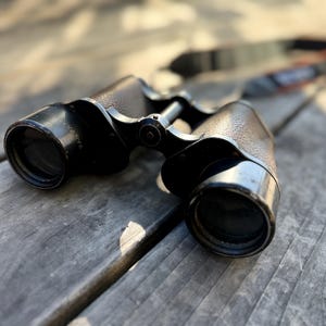 May include: Close-up of a pair of black binoculars with textured brown leather accents. The binoculars are resting on a weathered wooden surface, with a strap visible in the background. The lenses are dark and reflective.