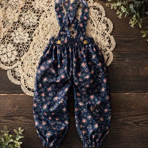 May include: A pair of navy blue overalls featuring a small floral pattern with pink and white flowers. The garment has an elastic waistband, two gold-toned buttons at the front, and shoulder straps that cross over the back. The pant legs have elastic cuffs.
